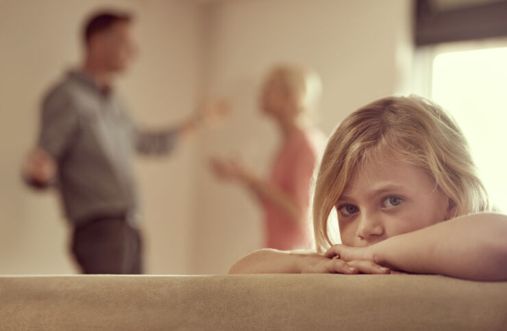 Liten flicka sitter i soffa med föräldrar som bråkar i bakgrunden