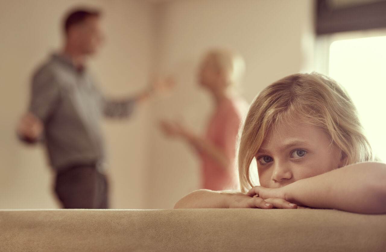 Little girl sitting on couch with parents arguing in the background.