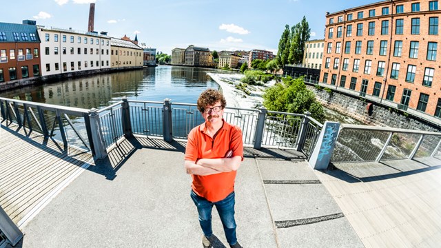 Man (Lars Stjernkvist) på bro med vatten och byggnader i bakgrunden