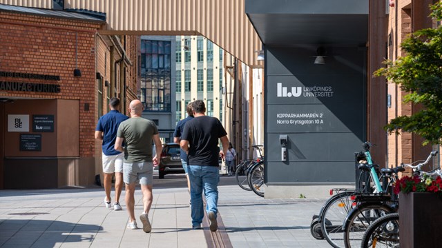 Men with their backs against the camera walk between buildings. Sign: LiU Linköpings universitet Kopparhammaren 2