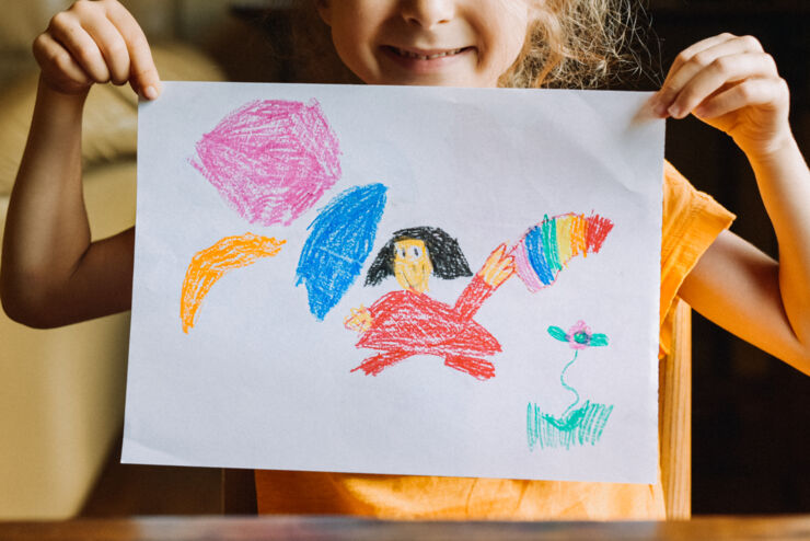 A girl is holding a drawing with colorful figures in front of her face.