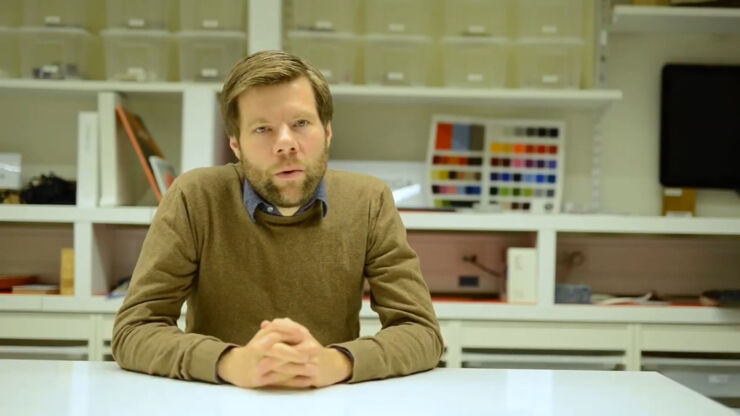 Man with beard and beige sweater sitting at a white table with a wall shelf in the background