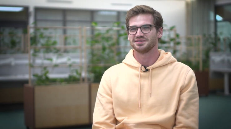 Young man wearing glasses and yellow hoodie with green plants in the background