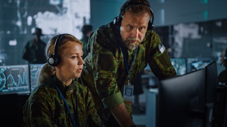 Woman and man with headsets and green military clothing in front of computer. More screens in the background.