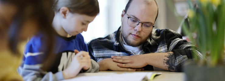 A student sits at his school desk and is helped by a teacher squatting next to him.