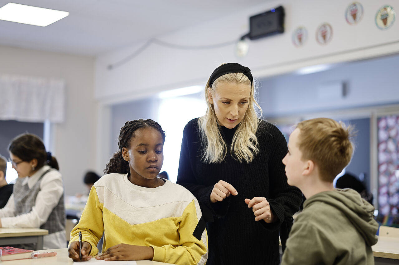 Lärare står och pratar med två elever som sitter vid ett bord