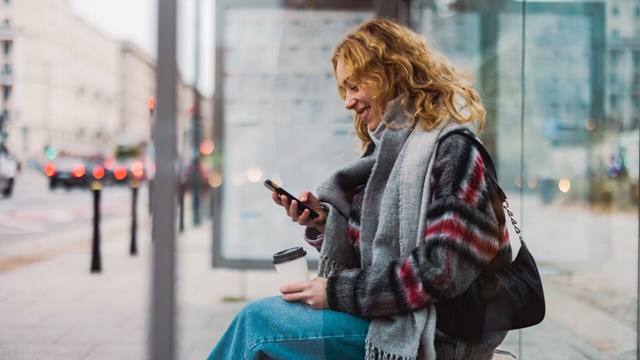 Kvinna sitter med sin smartphone vid en busshållplats.