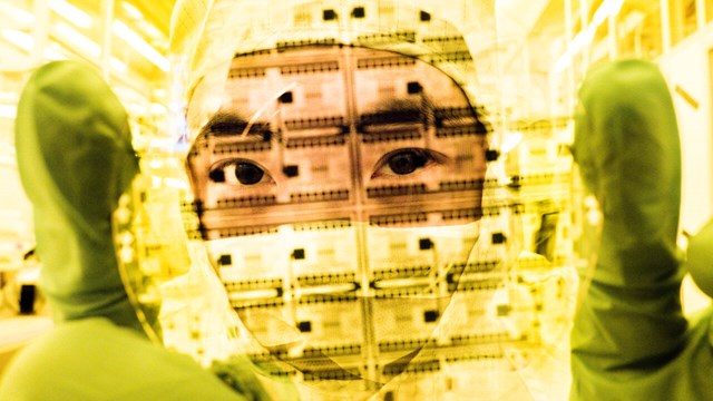 Man i heltäckande labbkläder håller upp en transparent skiva framför ansiktet.