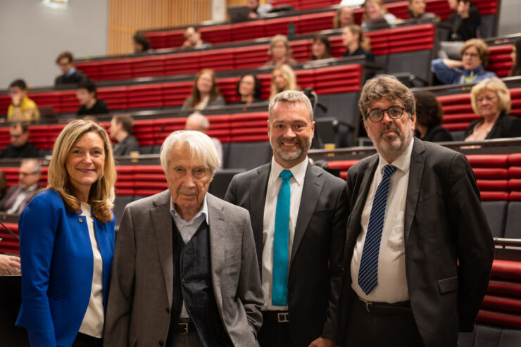 Tre män i kostym samt kvinna i blå klänning i föreläsningssal