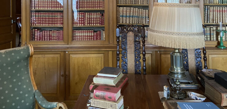 Bild inifrån ett slottsbibliotek. En bokhylla med gamla böcker täcker väggen, nedanför står ett bord med en äldre lampa och gamla böcker staplade på varandra.
