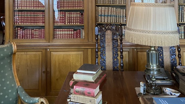 Bild inifrån ett slottsbibliotek. En bokhylla med gamla böcker täcker väggen, nedanför står ett bord med en äldre lampa och gamla böcker staplade på varandra.