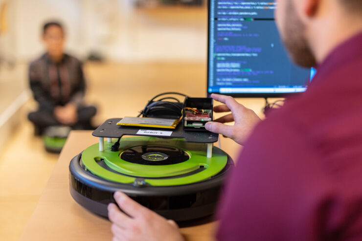 A person sits in front of a computer screen and handles a floor robot