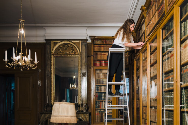En kvinna står på en stege vid en bokhylla och bläddrar i en bok.