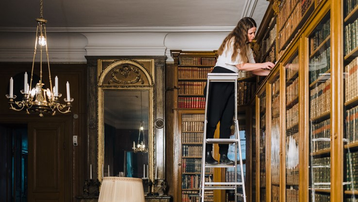 En kvinna står på en stege vid en bokhylla och bläddrar i en bok.