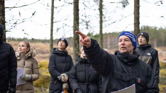 En person står och pekar ute i skogen med en lyssnande publik.