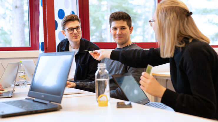 Kvinnlig student räcker fram ett kort till två manliga studenter. Uppfällda laptops och vattenflaskor på bordet.