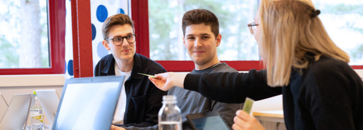 Kvinnlig student räcker fram ett kort till två manliga studenter. Uppfällda laptops och vattenflaskor på bordet.