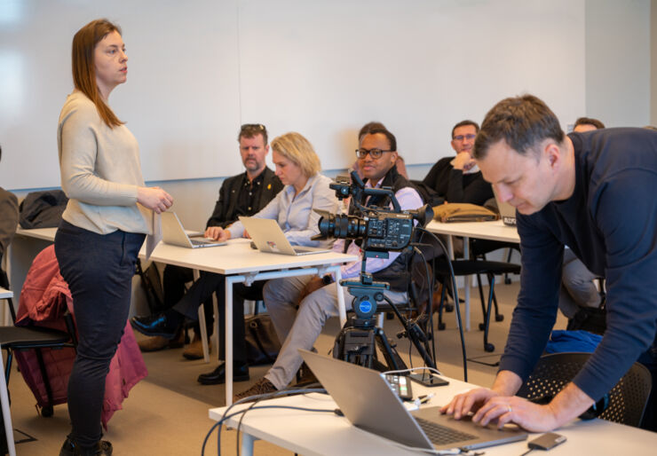 A woman stands in front of audience and talks. 