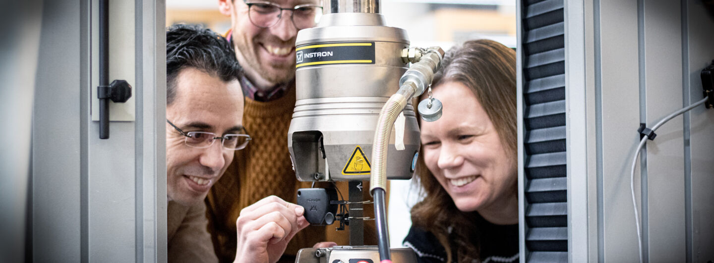 Three researchers standing by a machine that tests strength: 