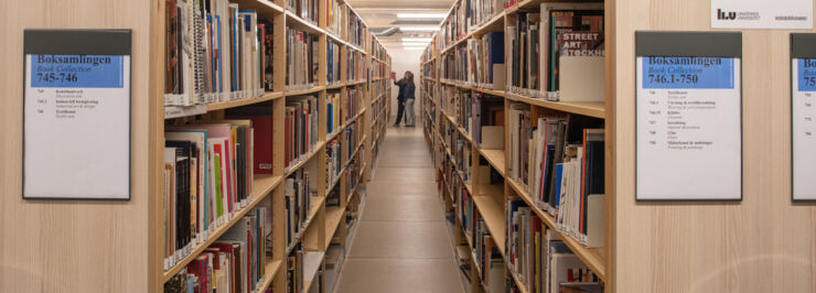 Two rows of book shelves and two people at one of the shelves in the background.