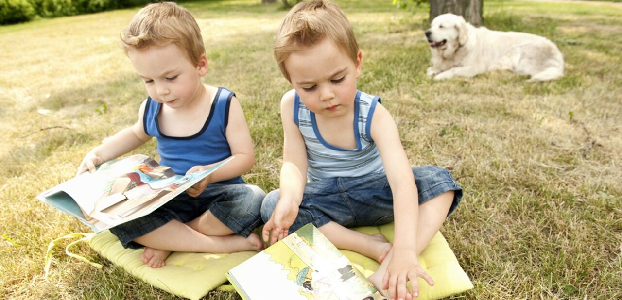 Tvillingpojkar som sitter på en filt i en trädgård och läser böcker.
