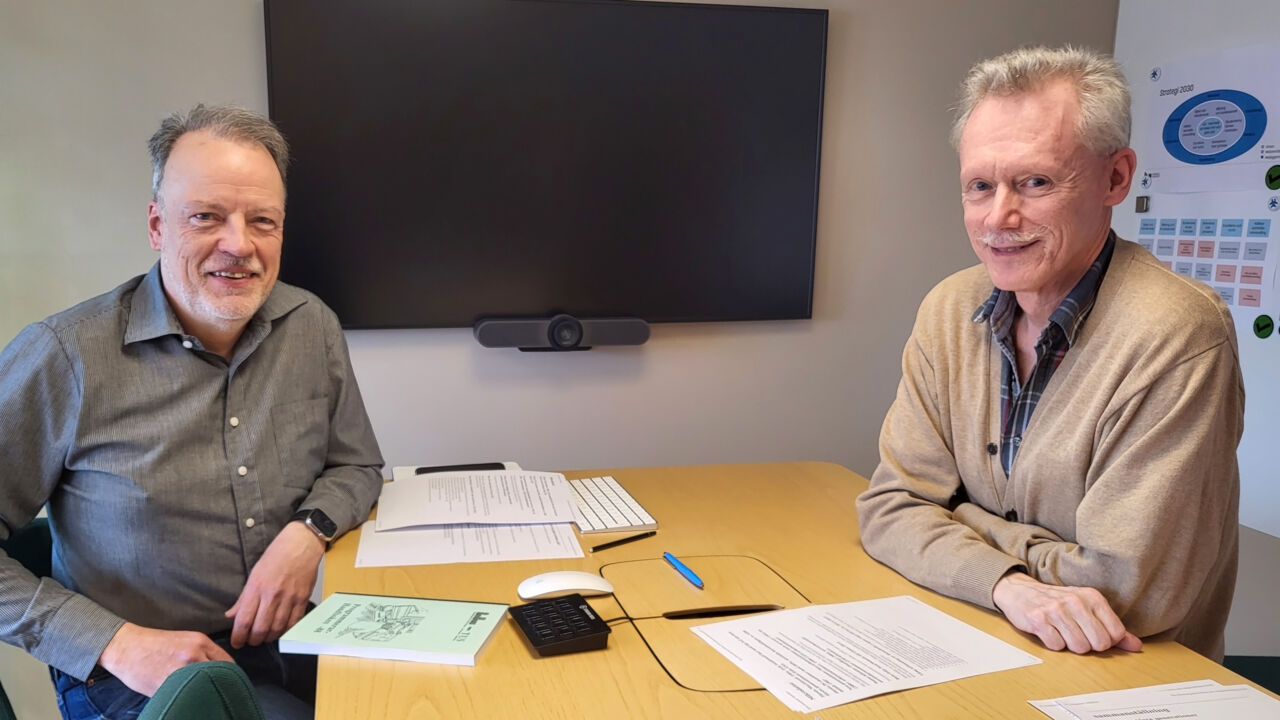 Två personer sitter på var sin sida om ett bord med diverse papper framför sig.