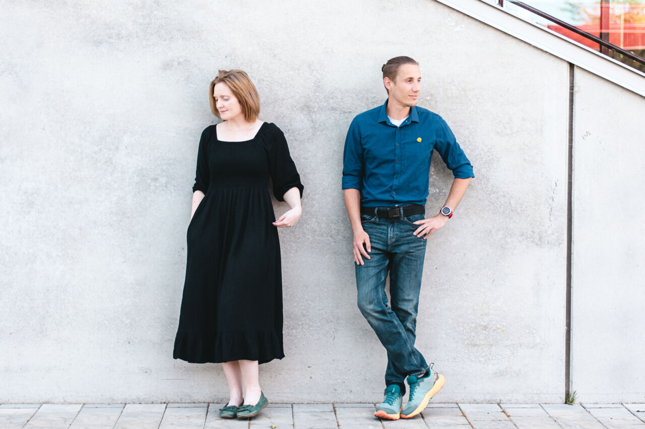 Alice Langley and Jean-Pierre Roux is standing by a concrete wall.