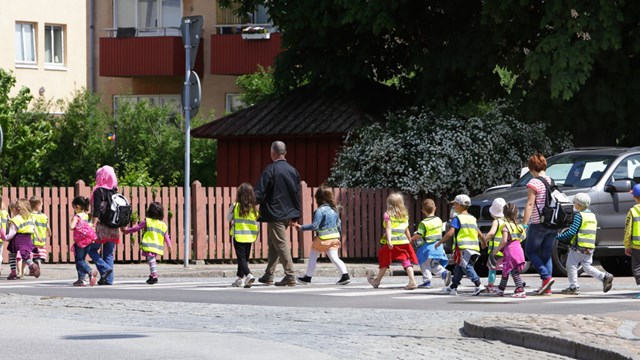 A group of children in yellow reflective vests are crossing the street together with some adults.
