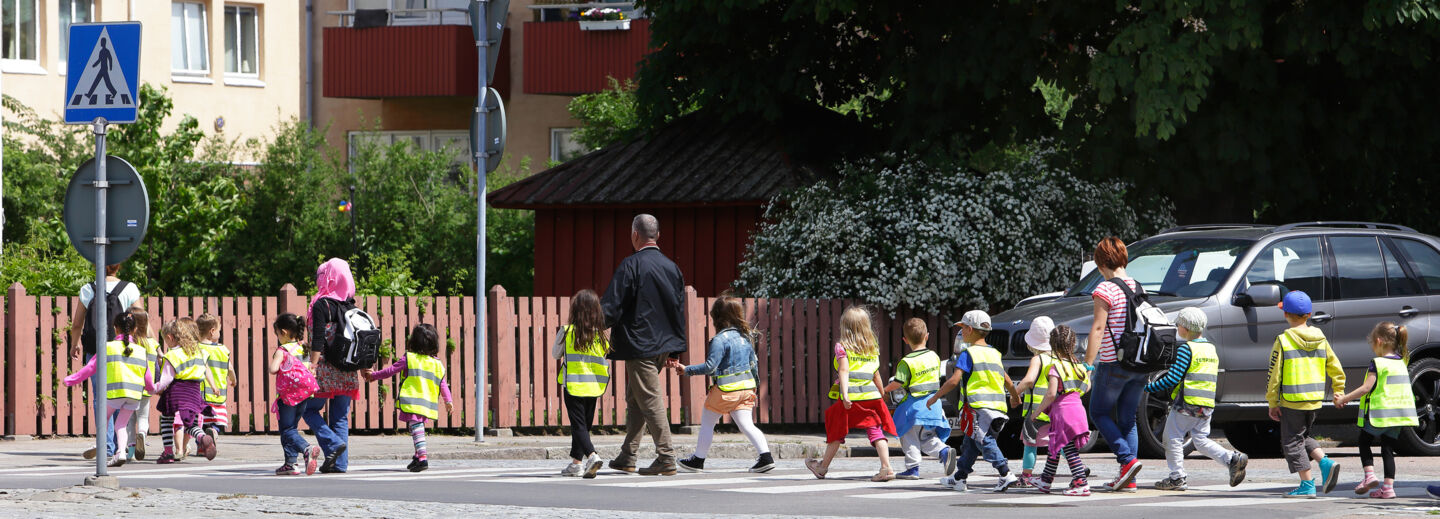 A group of children in yellow reflective vests are crossing the street together with some adults.