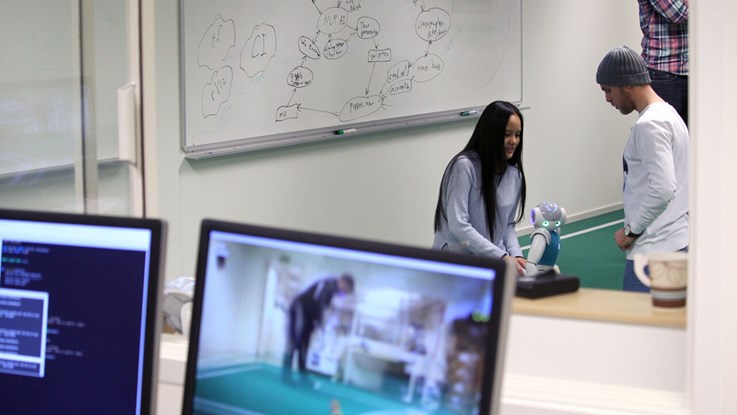 Två studenter håller på med en robot, datorskärmar i förgrunden