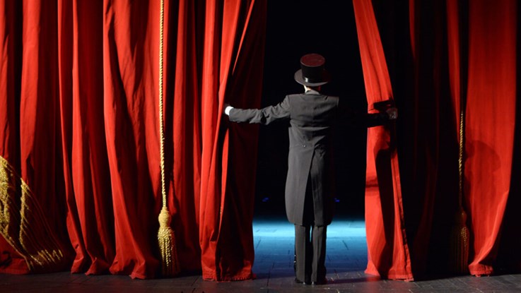 A man in a tuxedo opens a red curtain.