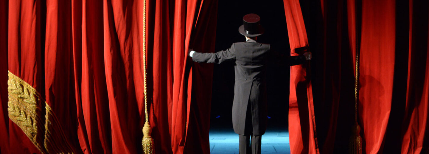 A man in a tuxedo opens a red curtain.