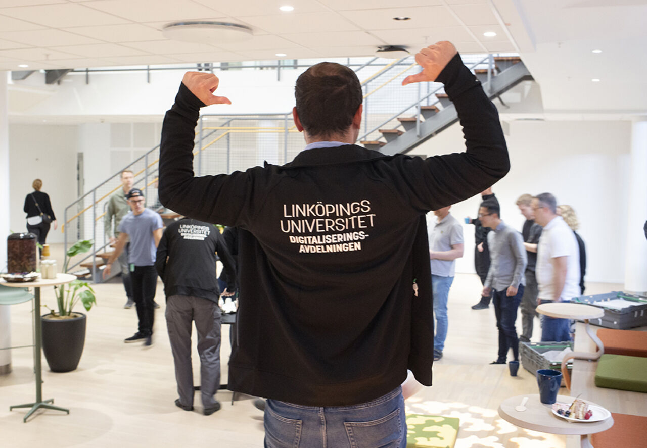 En man står med ryggen mot kameran, med armarna lyfta i vinkel. Tummarna pekar ner mot hans rygg. Texten på den svarta tröjans rygg lyder: Linköpings universitet, Digitaliseringsavdelningen.