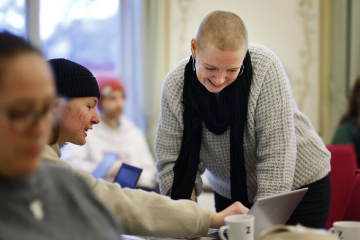 Kvinna undervisar vuxna elever. Läraren har en grå tröja, svart halsduk och rakat hår.