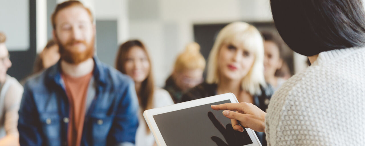 En kvinna med en ipad sitter framför en publik som lyssnar på henne. 