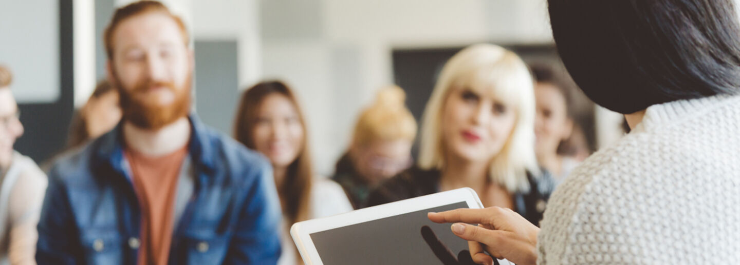 A woman with an iPad sits in front of an audience listening to her.