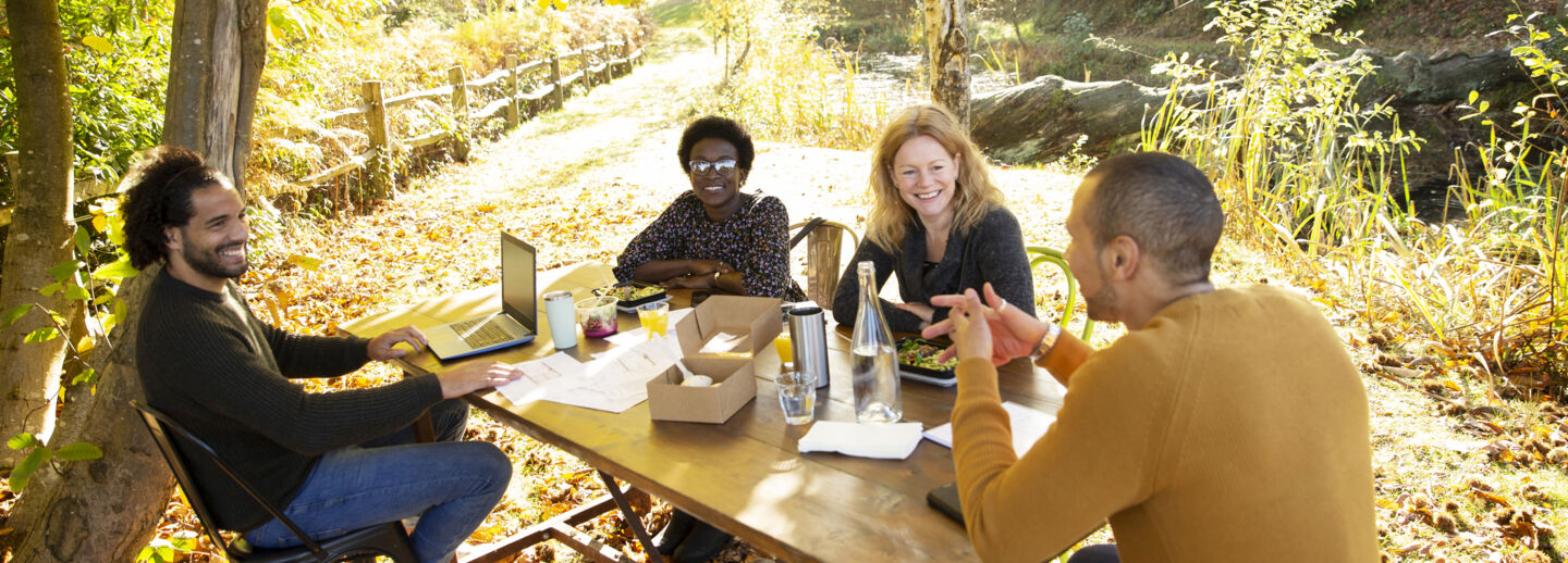Four People sitting at a picknick table outside in the woods. There is a laptop and coffee mugs on the table.