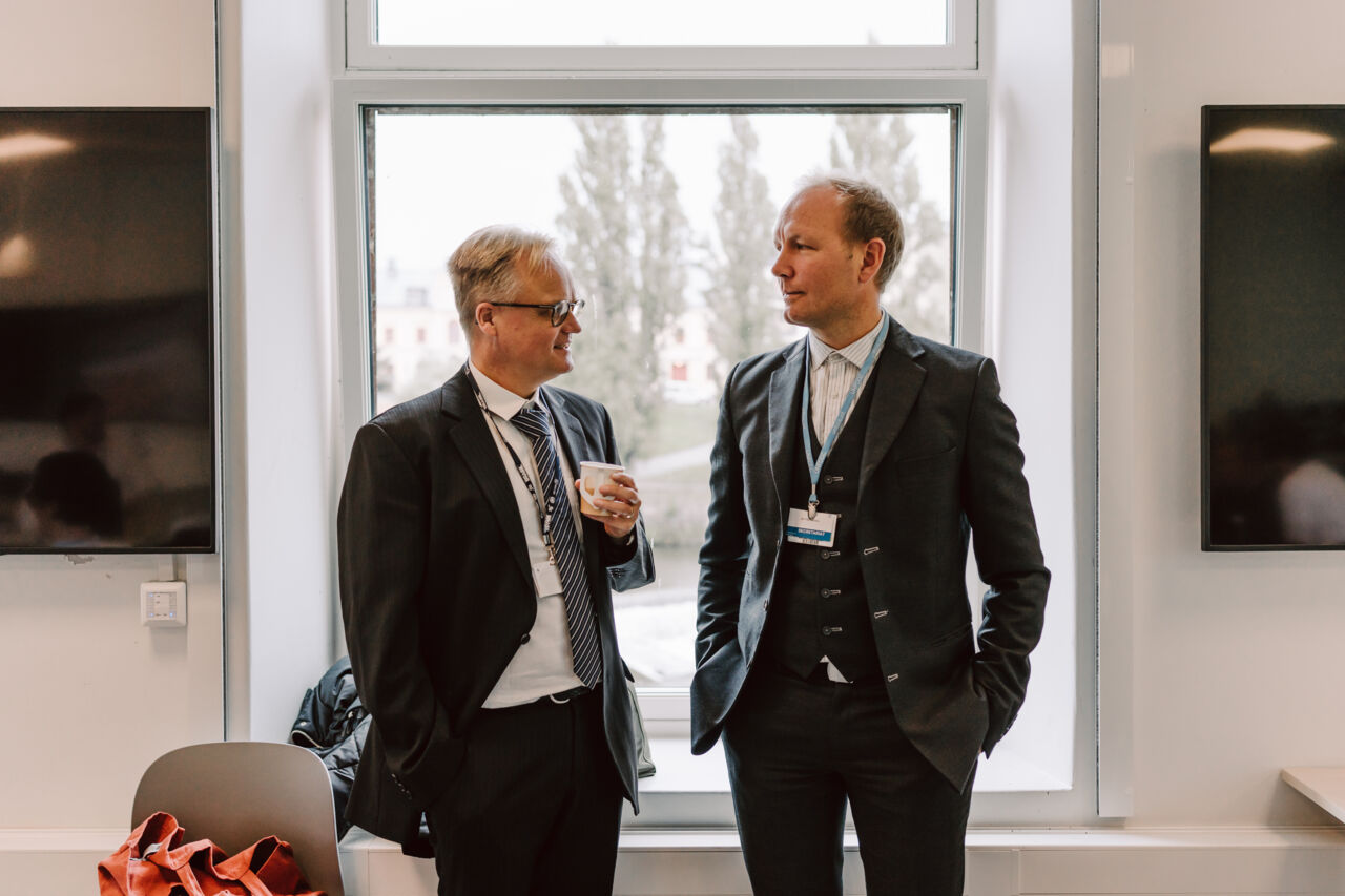 Två lärare i kostym står och pratar med varandra framför ett fönster