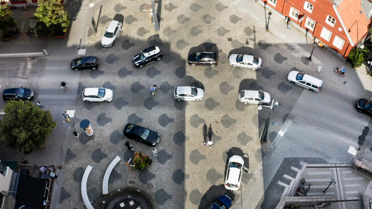 A four-way intersection from above where seven black and seven white cars meet. Cyclists and pedestrians can also be seen at the intersection. 