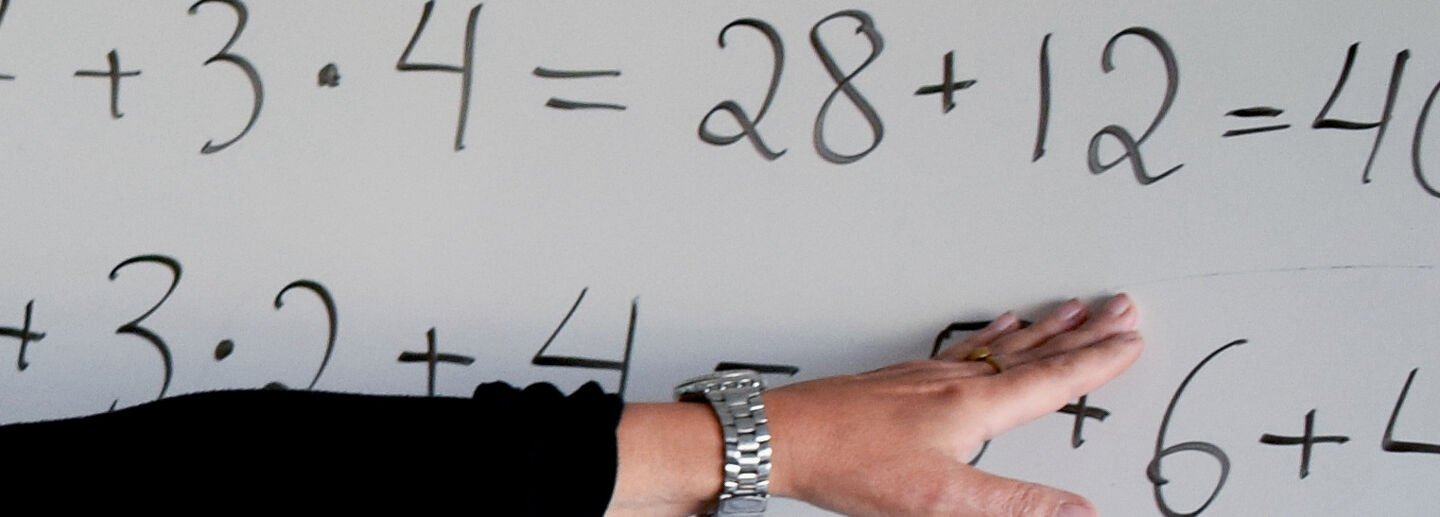 An arm wearing a black shirt and a watch is pointing to math problems written with black pen on a whiteboard 