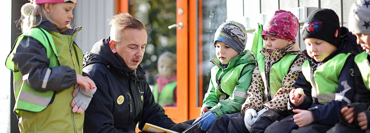 En lärare sitter på huk utomhus och håller i en bok. Runt läraren sitter barn i vinterkläder och lyssnar till läraren. 