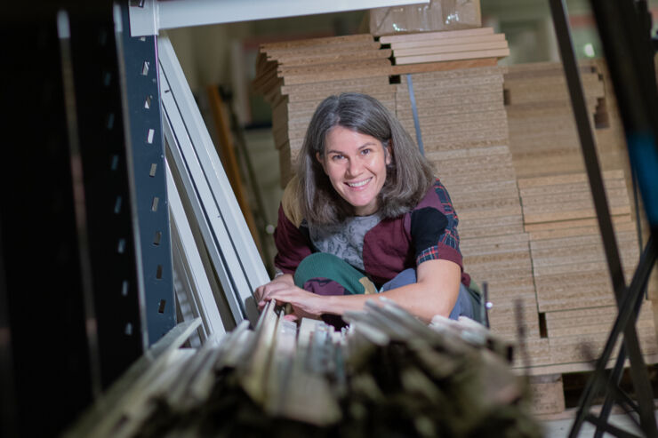 Smiling woman peeking out from behind a pile of wooden boards.