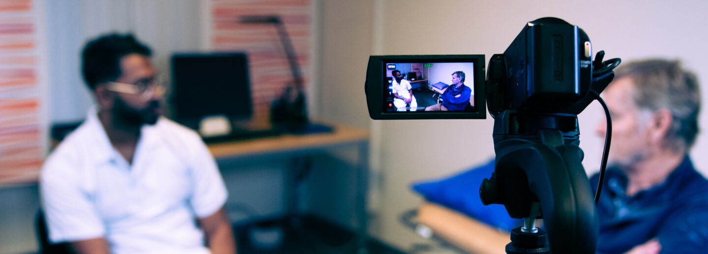 Två personer samtalar i ett rum, fotade genom en videokamera som syns i förgrunden.