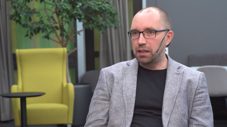 Man in grey jacket and glasses with yellow armchair and plant in the background