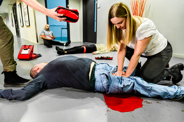 Demonstration av skadeplats, en kvinna trycker händerna mot benet på en liggande person, fiktivt blod illustrerar blödning.