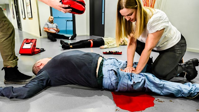 Demonstration av skadeplats, en kvinna trycker händerna mot benet på en liggande person, fiktivt blod illustrerar blödning.