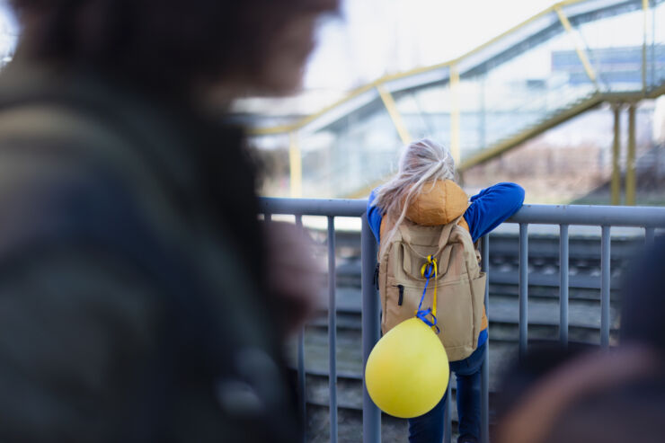 Flicka som står vid ett staket framför järnvägsspår. Hon bär en ryggsäck som har en gul ballong fastknuten med blågula band.