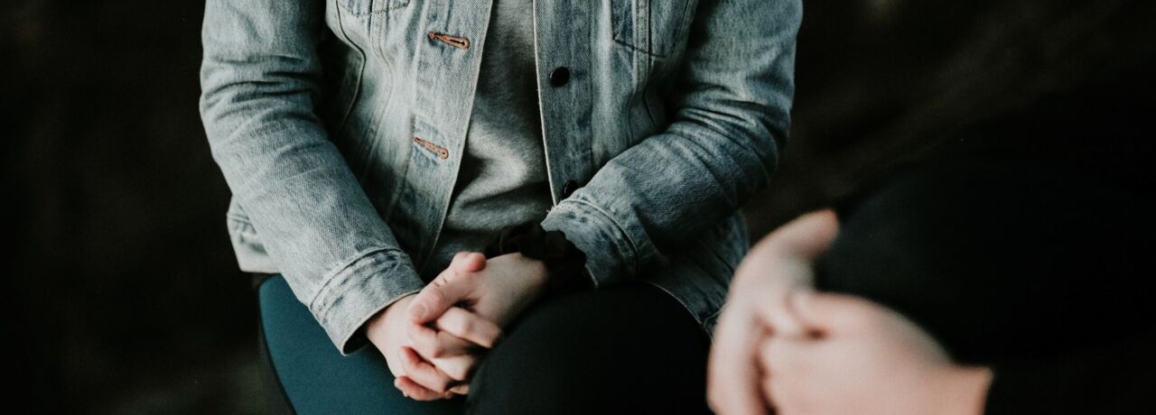 A person in a denim jacket with his hands on his knees sits opposite someone who is gesturing with his hands.