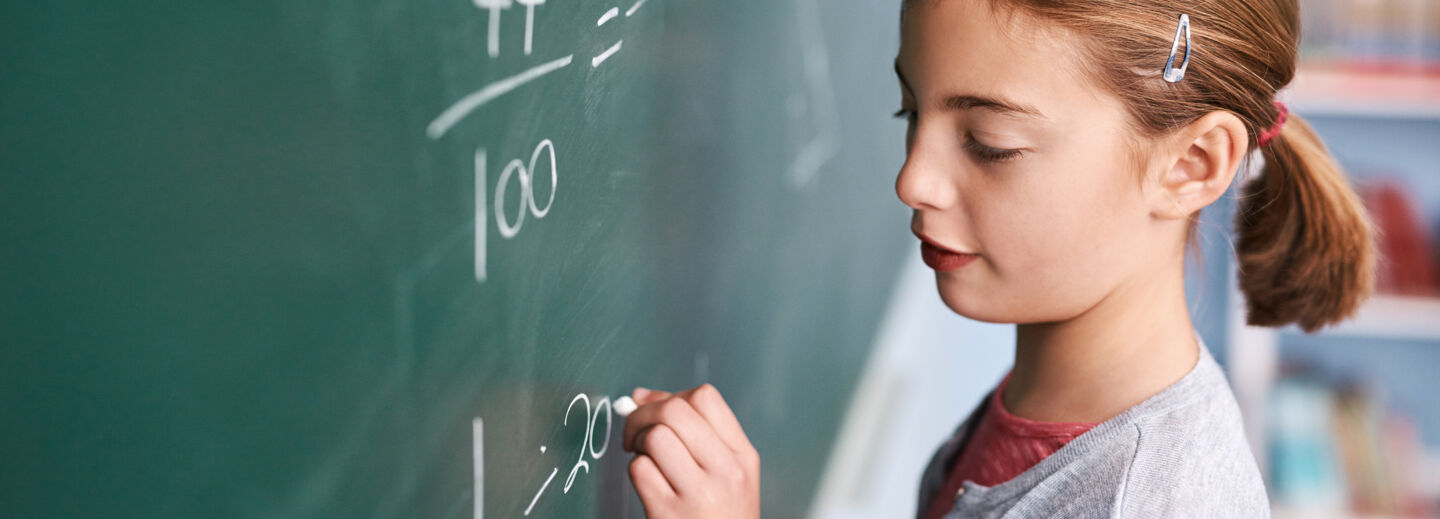 Flicka i brunt hår står framför en skrivtavla i klassrummet och räknar matte