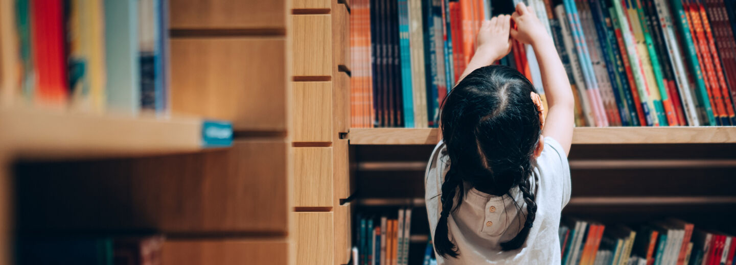 Girl reaches for a book on a bookshelf.